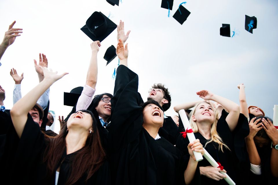 A group of graduates in black gowns joyfully throw their caps into the air while holding diplomas.