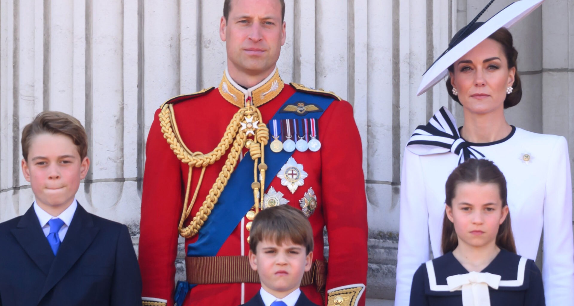 Prince William wears red military uniform and Kate Middleton wears a white dress with navy detailing, as they stand on the Buckingham Palace with their three kids, Prince George, Prince Louis, and Princess Charlotte