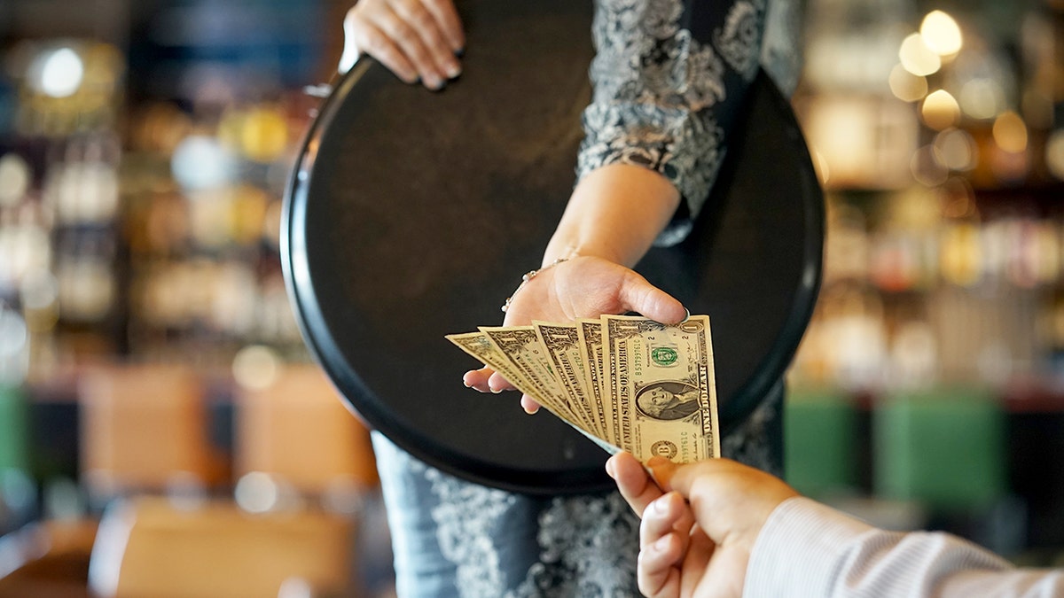 A hand holding $5 in cash is given to a restaurant server as a tip.