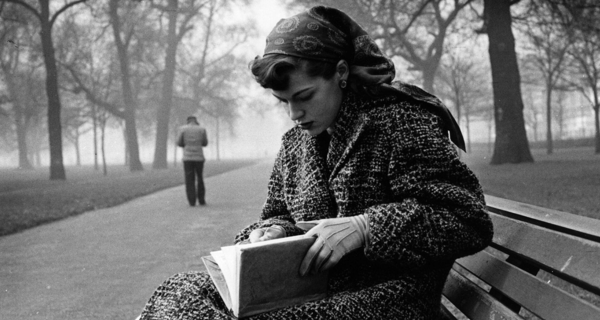 Black and white image of a woman wearing a winter coat and headscarf, reading a book on a park bench