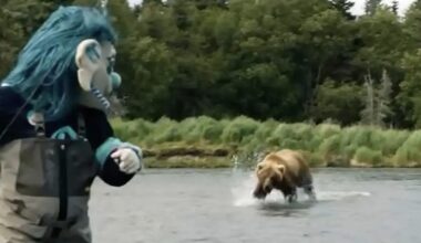 A person in a blue troll costume stands in shallow water, looking at a brown bear running through the water nearby. Green trees and grass line the background.