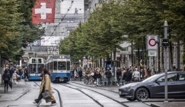 Pedestrians cross tram tracks on a busy shopping street in Zurich as trams and cars wait, with a Swiss flag overhead.
