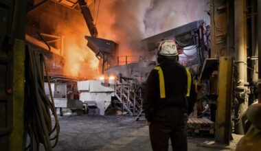 An employee wearing a helmet and safety gear observes an electric arc furnace emitting bright orange flames and smoke inside the Liberty Steel Aldewerke mill.