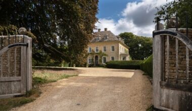 Dean Manor, a large stone house with symmetrical windows and ivy on the facade, is viewed through open wooden gates.