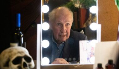 Nick Ullett, the actor and writer, is reflected in a dressing room mirror, lights around it, and a skull to his left.