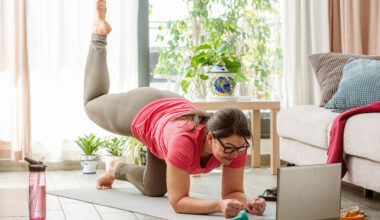A woman practices Pilates at home in her living room on a mat. She is balancing, close to the ground, on her forearms and her bent left knee. Her right leg is bent at 90° and elevated behind her. In the room we see a wtaer bottle, a couch, a plant and a coffee table. A laptop is open in front of her.