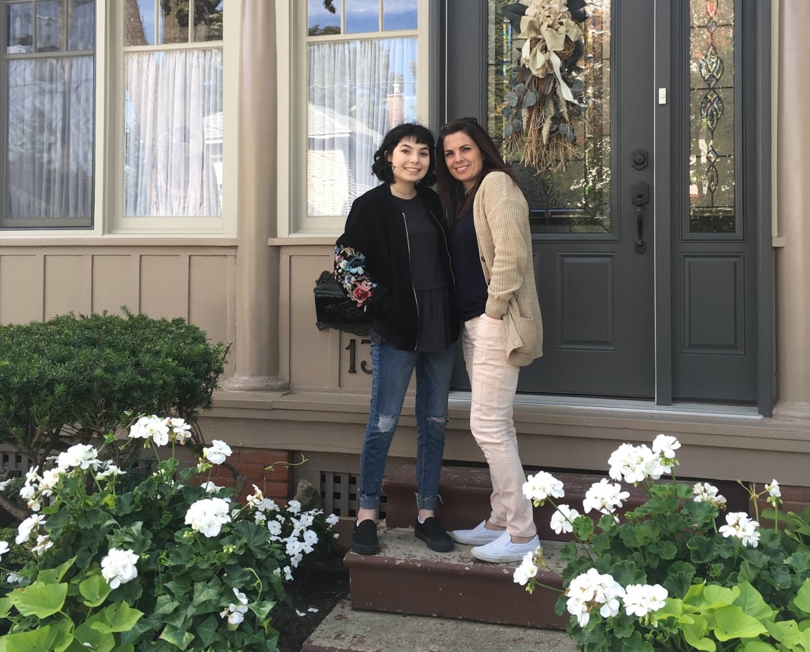 Two smiling women stand on a front porch and pose for a photo. 