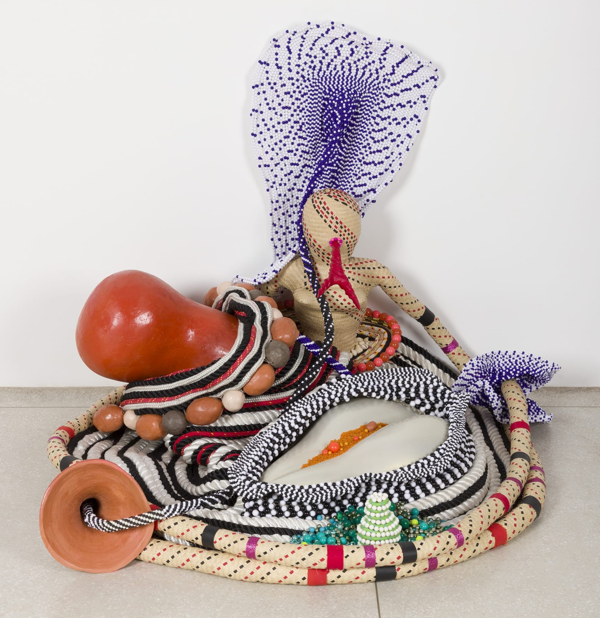 an abstract sculpture that sits on the floor and leans on the wall, with beads, wood, straw, resin, and other materials