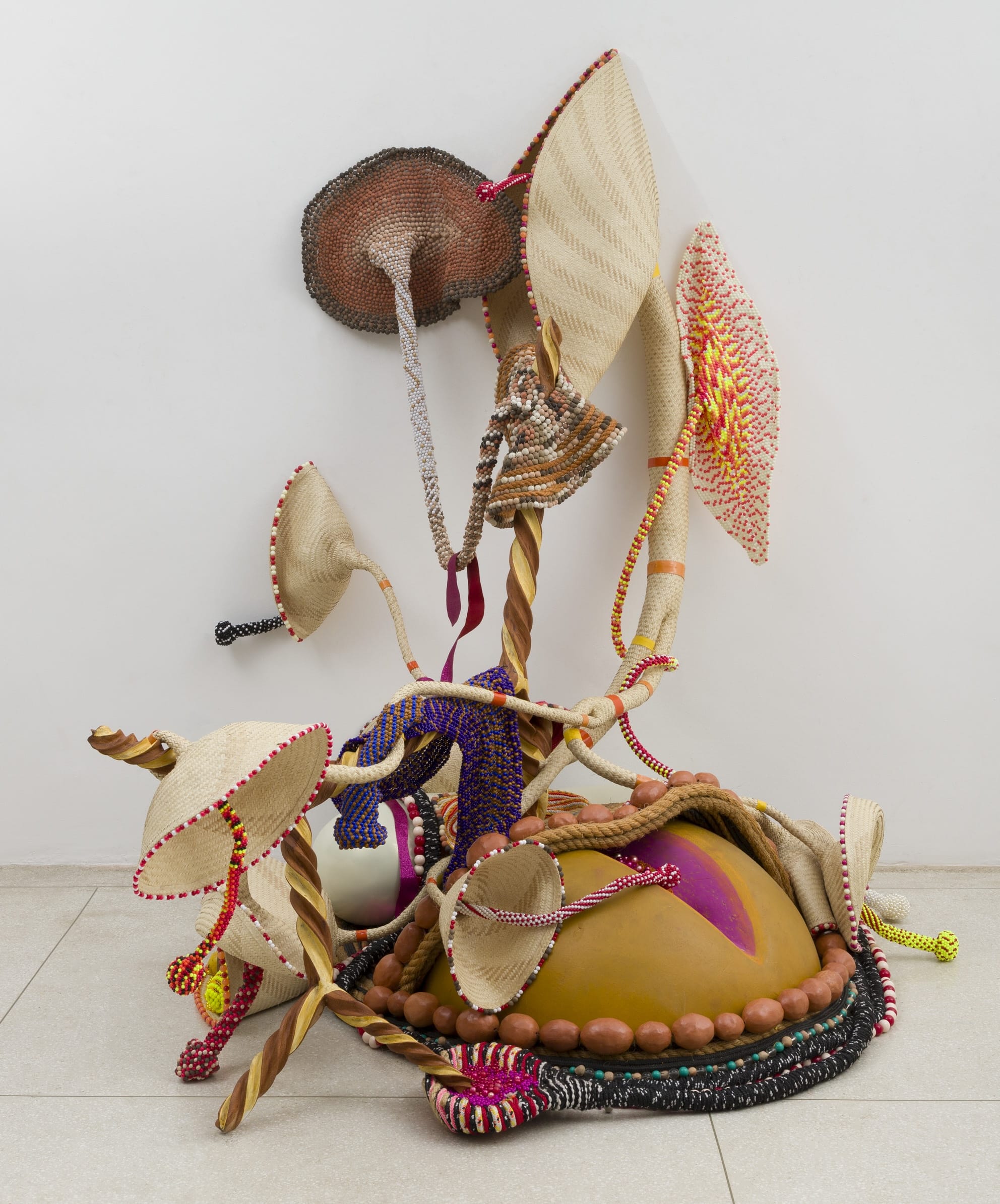 an abstract sculpture by Maria Nepomuceno that sits on the floor and leans on the wall, with beads, wood, straw, resin, and other materials