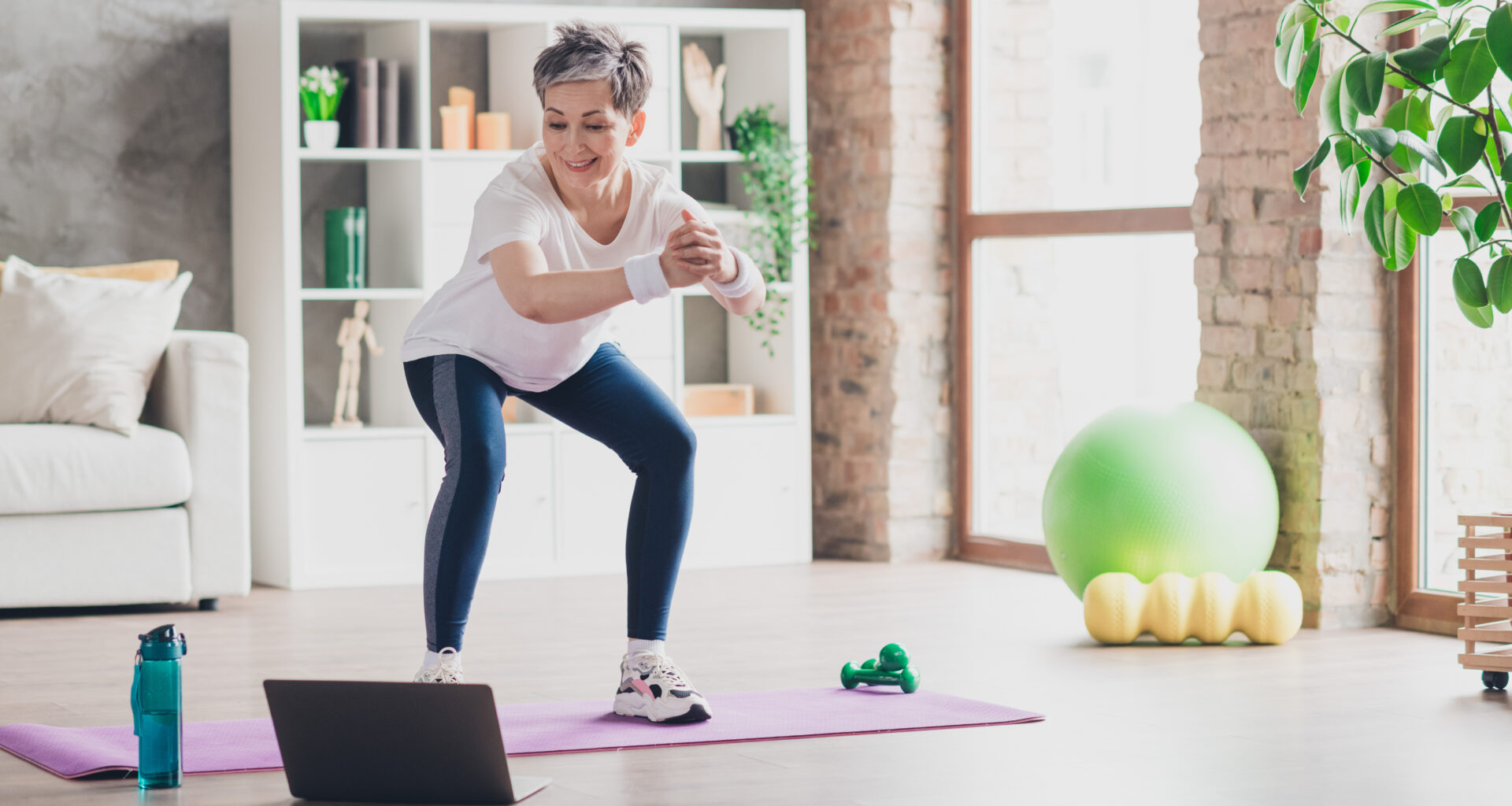 A woman performs a bodyweight squat at home on an exercise mat, while watching an open laptop at her feet. We see a water bottle, light dumbbells, an exercise ball and a foam roller in the room, along with a couch and bookshelves.