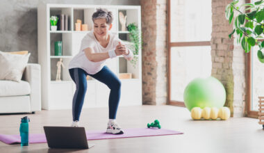 A woman performs a bodyweight squat at home on an exercise mat, while watching an open laptop at her feet. We see a water bottle, light dumbbells, an exercise ball and a foam roller in the room, along with a couch and bookshelves.