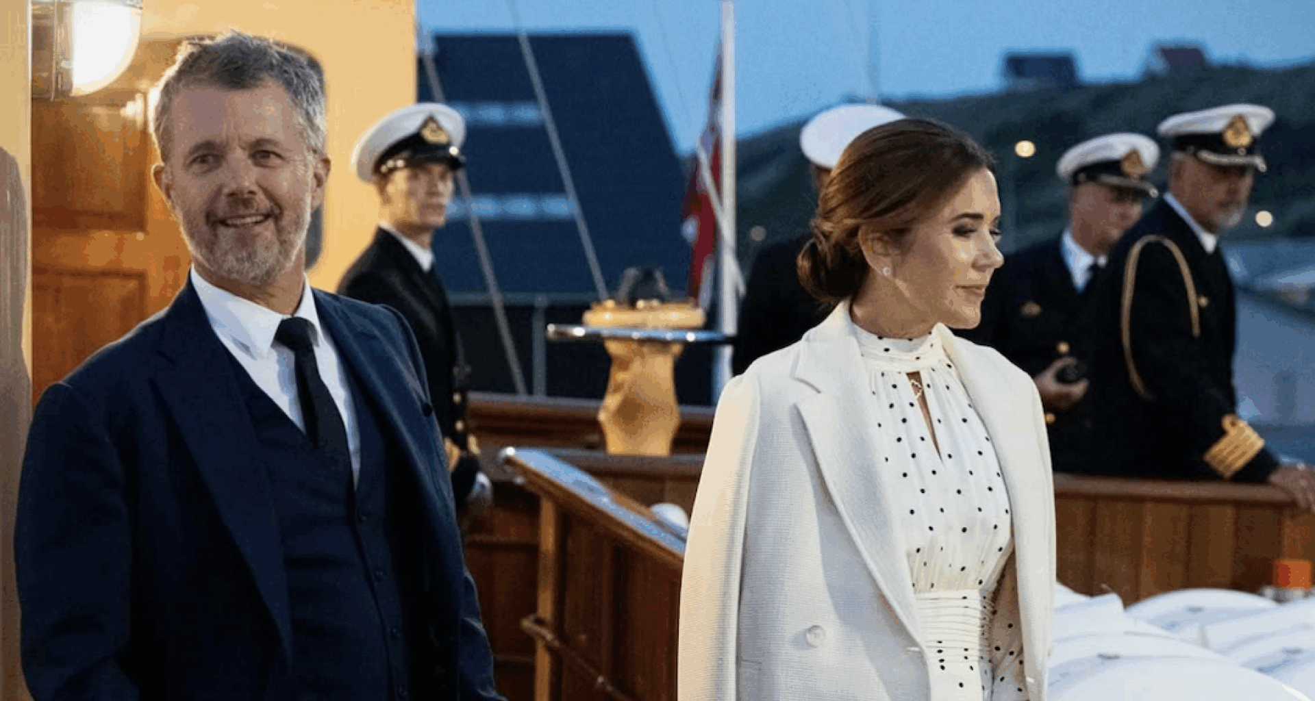Queen Mary wearing a white polka dot dress and King Frederik in a suit standing on the royal yacht