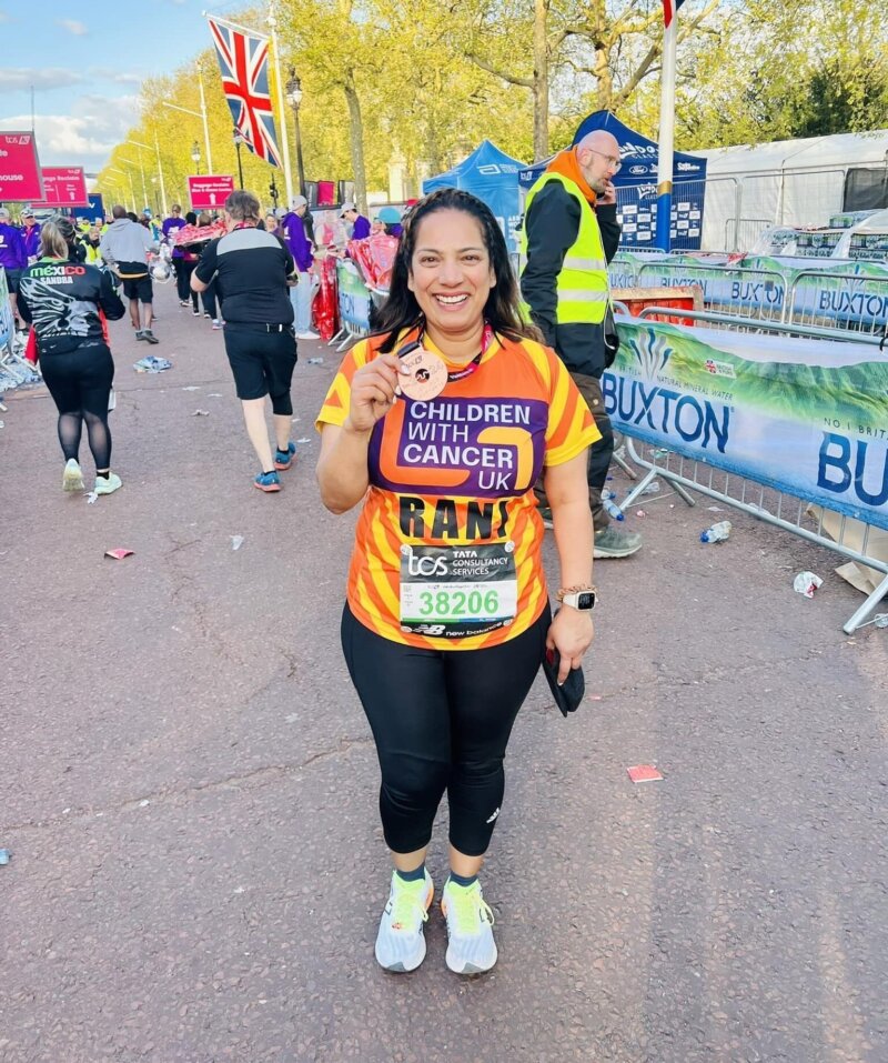 A middle aged woman is standing at the finish line of a long race, wearing sporty clothes and holding up a medal for the camera. She looks tired but proud. It's a sunny day.