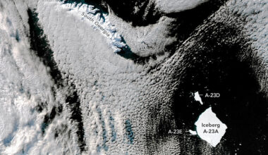 A large, white, square-shaped iceberg floats amid black water in the bottom right of the satellite image. Two smaller icebergs flank the iceberg on its left and top-left sides. Crescent-shaped South Georgia, an island covered in snow and ice, is toward the top-left. White clouds cover a sizable part of the ocean.