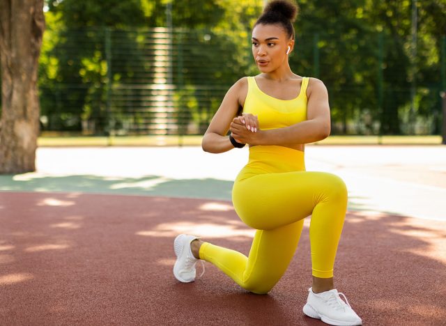 Sport And Healthy Lifestyle. Fit black woman doing curtsy lunges with twist, exercising outdoors on basketball stadium. Portrait of confident lady training in wireless earbuds, full body length