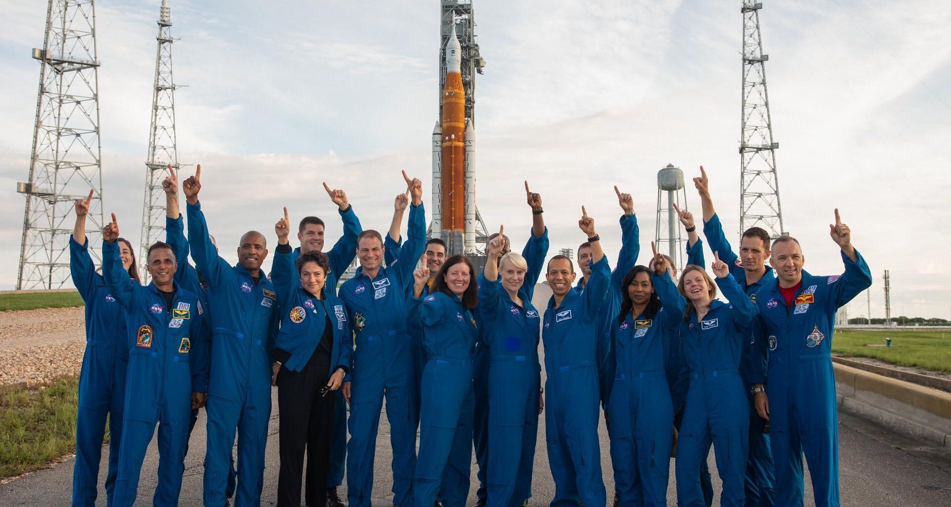 a bunch of astronauts in flight suits standing in front of a huge rocket and pointing at the sky