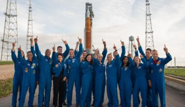 a bunch of astronauts in flight suits standing in front of a huge rocket and pointing at the sky