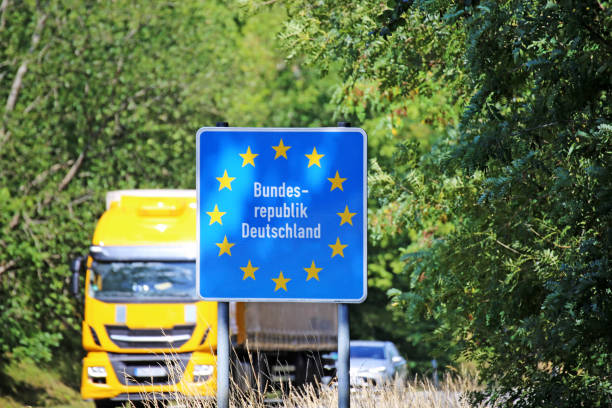 Blue Germany border sign with EU stars, in front of a yellow truck and cars on a road surrounded by trees.