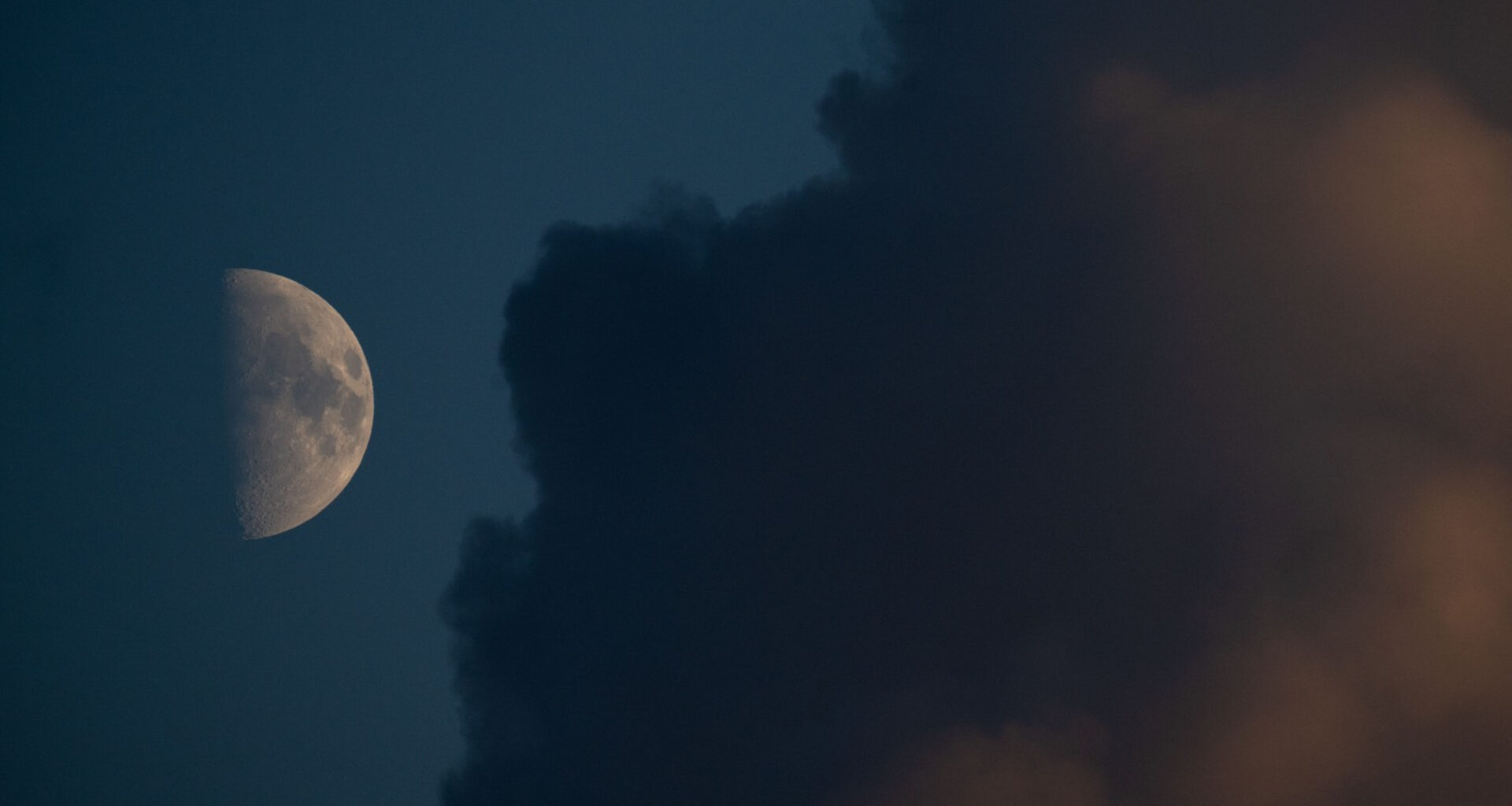 An image of a half-lit moon against a dark blue sky, with its right half lit by direct sunlight and its left shrouded in shadow. Dark plains called lunar maria can be seen covering swathes of the lit side and a large cloud dominates the right side of the image.