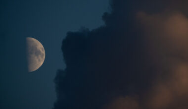 An image of a half-lit moon against a dark blue sky, with its right half lit by direct sunlight and its left shrouded in shadow. Dark plains called lunar maria can be seen covering swathes of the lit side and a large cloud dominates the right side of the image.
