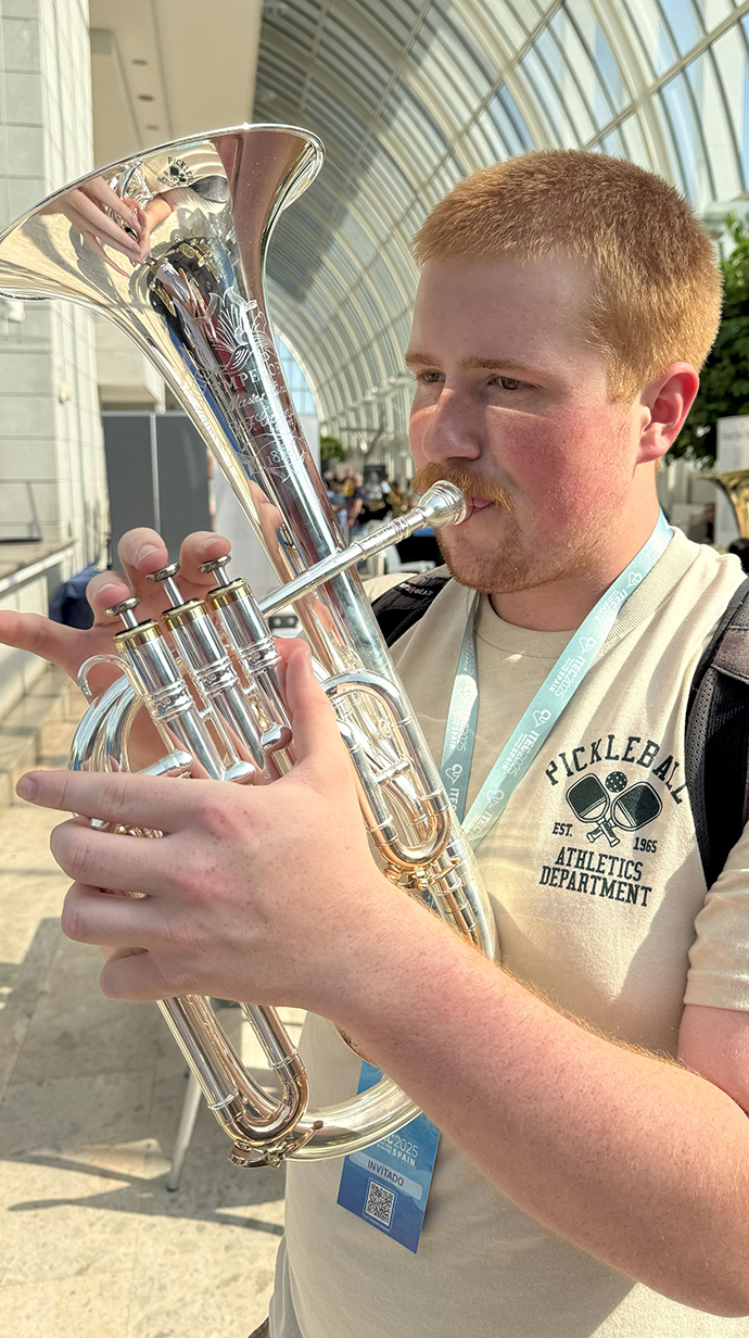 A young man plays a brass instrument in an atrium with a glass ceiling.
