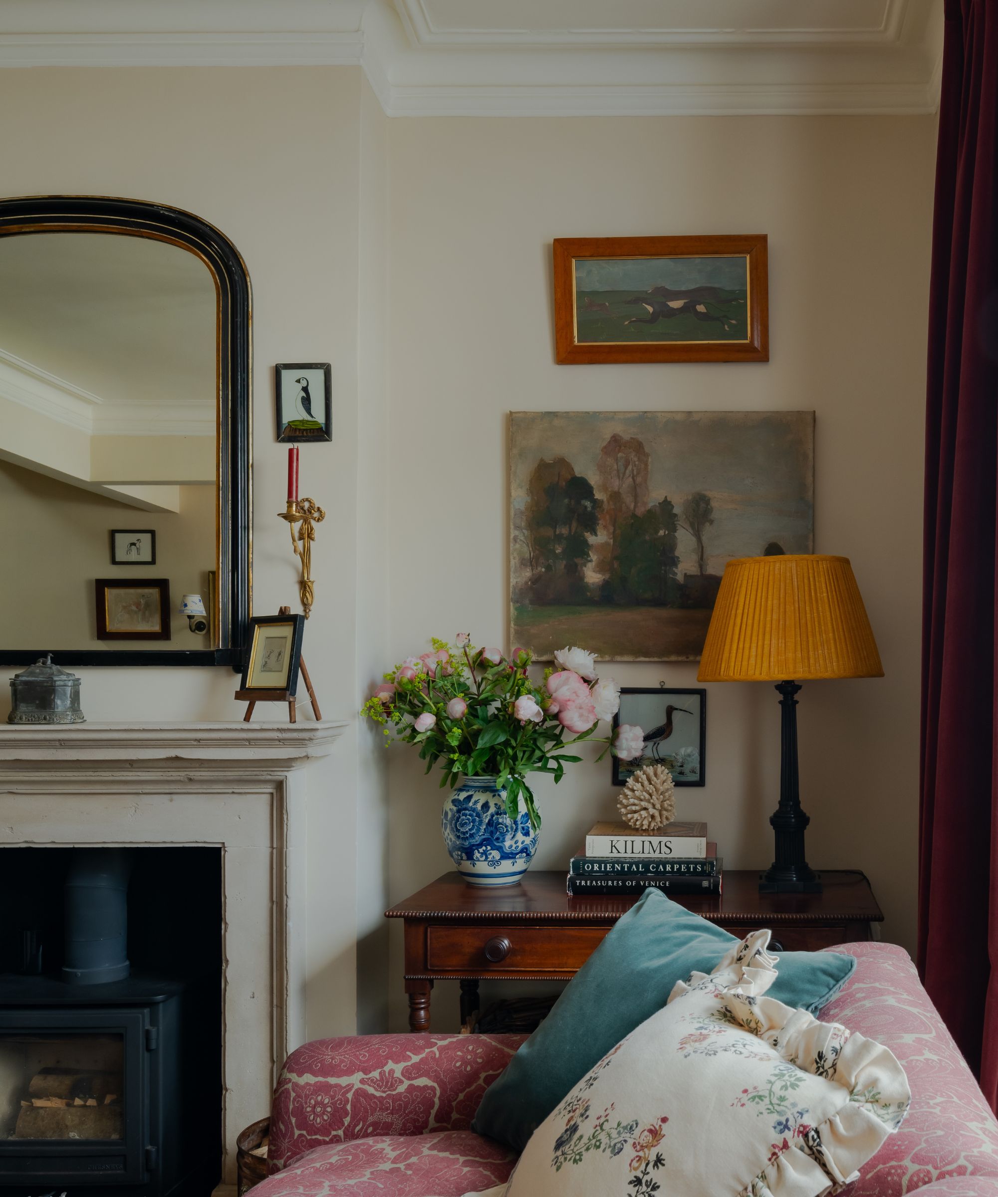 Living room with antique arched mirror and fireplace, vintage art on walls, pink paisley sofa with cushions and china vase with flowers