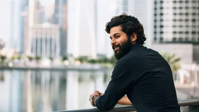 Young man with beard standing outside with a city backdrop, showing quiet wealth and luxury.