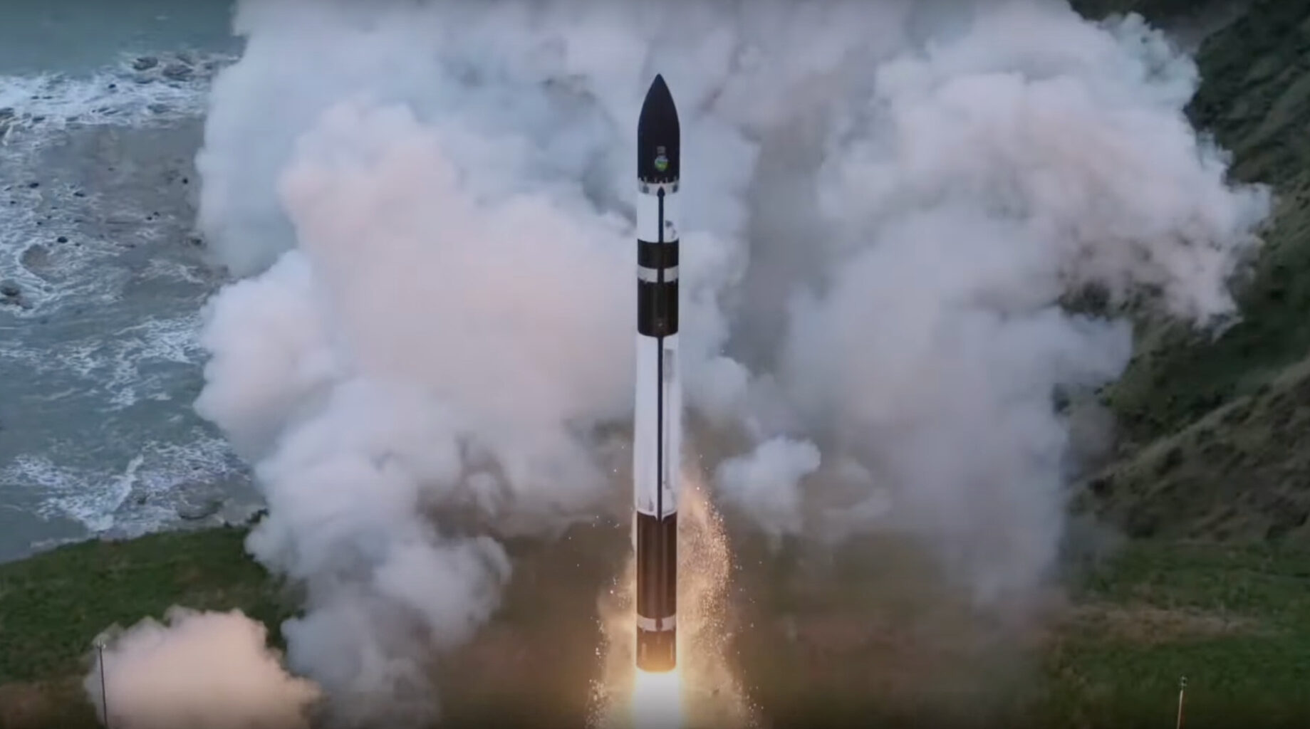 a black and white rocket rises off a seaside launch pad, with a plume of white exhaust in the background