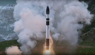 a black and white rocket rises off a seaside launch pad, with a plume of white exhaust in the background