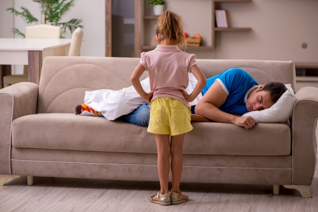 Young man sleeping on a couch while his young daughter watches him.