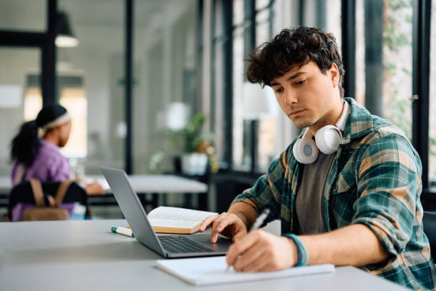 A student with headphones around his neck studies with a laptop and notebook at a desk in a classroom.