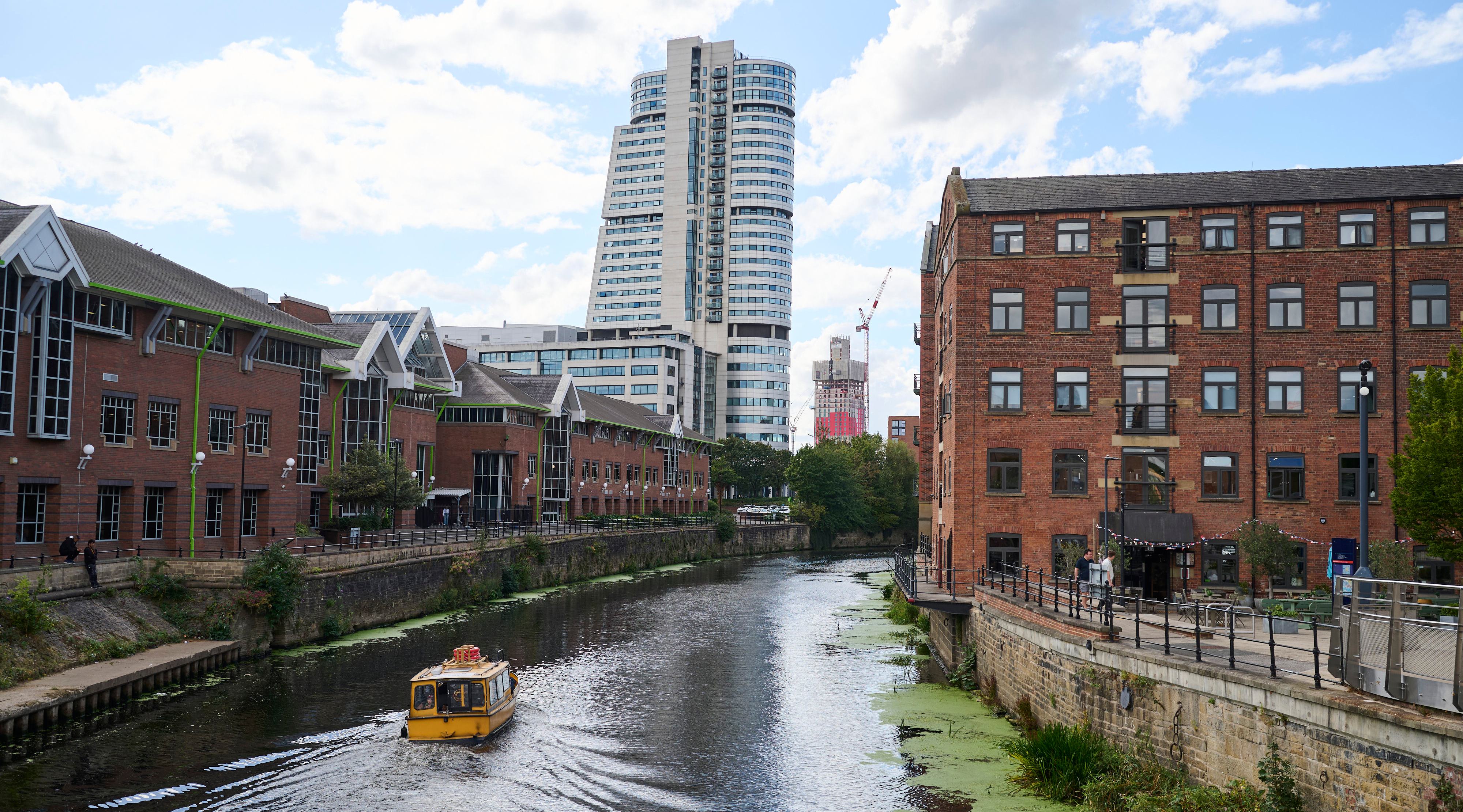 A yellow riverboat on the Leeds canal, with modern and older buildings in the background.