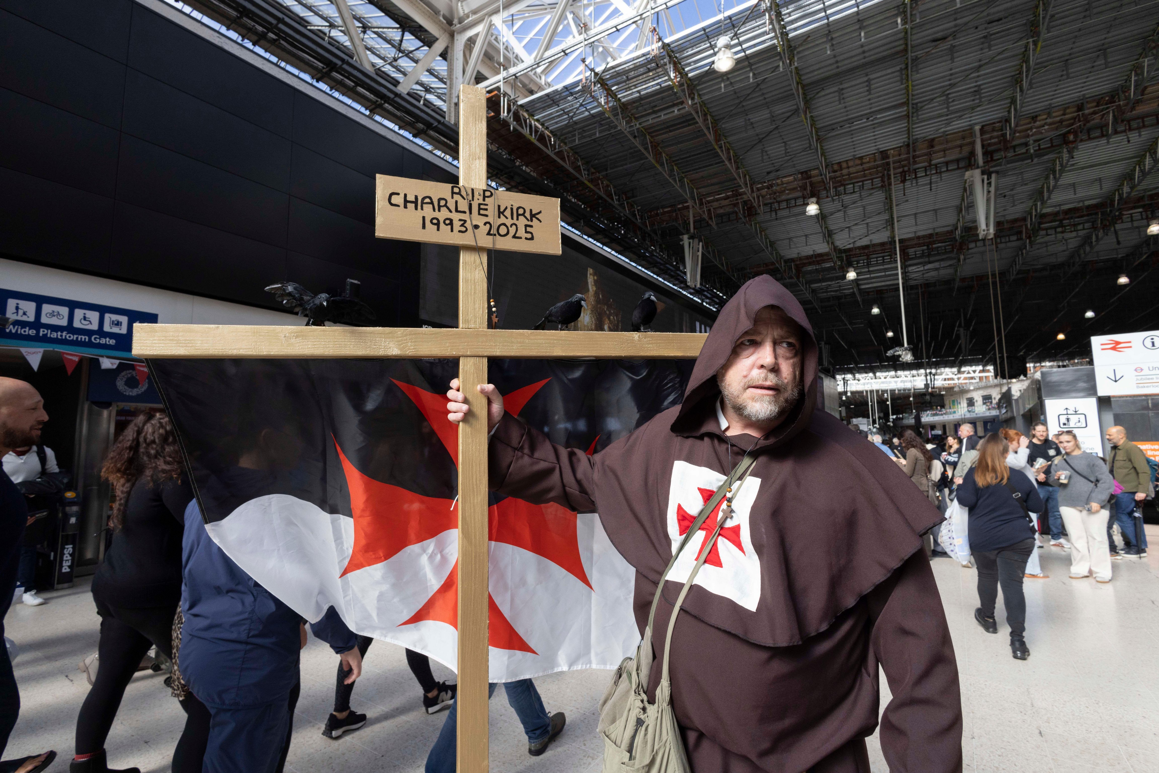 Person in monk's robes holding a cross with a Charlie Kirk memorial at Waterloo Station.