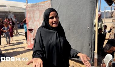 Sylvia al-Shurafi, a woman wearing all black, pictured in front of a make-shift divider constructed from beams and two large pieces of fabric with some tears in them. She has a serious expression and has her hands out in front of her. Behind her a group of people including children looks on.