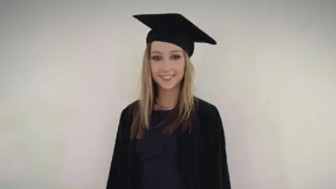 Family photo Jessica Gallagher smiling at the camera on her graduation day.  She is wearing a black mortarboard and a black robe over a black or dark navy dress.  She has long, straight blonde hair and is standing in front of a plain, pale background. 