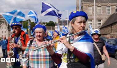 A woman draped in a saltire plays a flute at the front of a crowd of pro-independence marchers