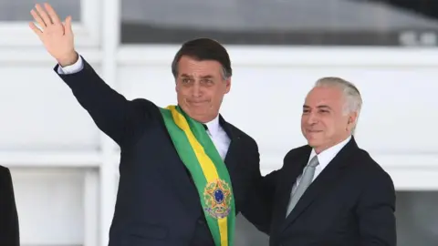 AFP via Getty Images Jair Bolsonaro, wearing the presidential sash over a black suit, waves to supporters during his swearing-in ceremony. Next to him is the outgoing Brazilian president, Michel Temer