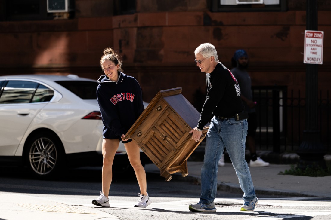 Here's alt text for this image:
A woman wearing a black "Boston" sweatshirt and a man in a black t-shirt and jeans work together to carry a wooden piece of furniture, likely a small dresser or nightstand with drawers. 