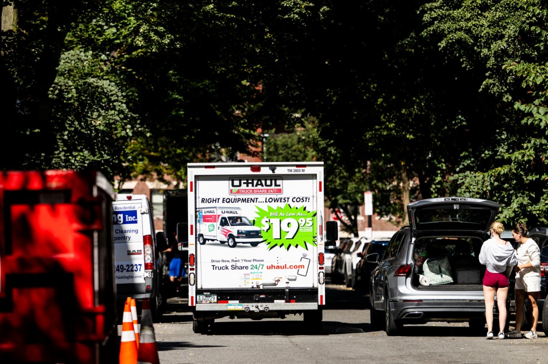 Students unloading belongings from the back of an SUV on a tree-lined street. A U-Haul rental truck is visible in the background with "$19.95" pricing advertised on its side.
