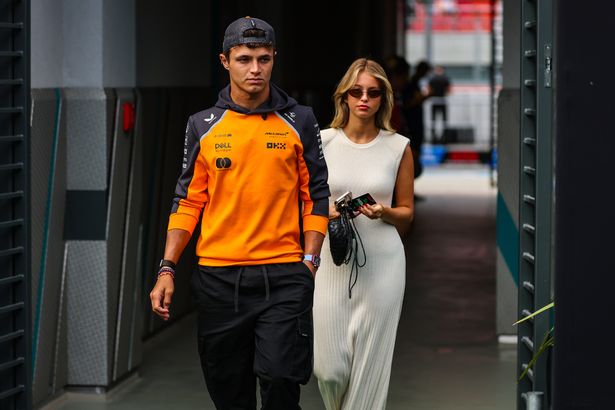 Margarida Corceiro, in a white dress, and Lando Norris, in an orange hoodie and dark trousers, walking into the paddock at a racetrack in August 2025.