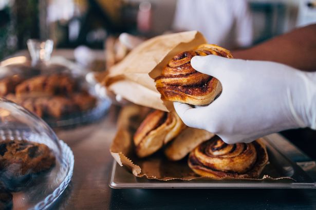 A close-up of a freshly baked cinnamon roll being served by a gloved hand in a bakery