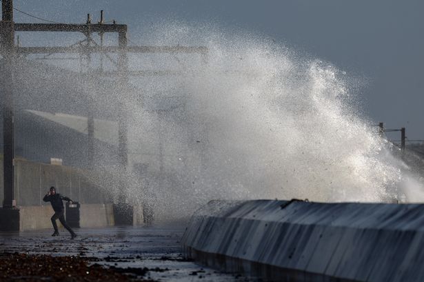Members of the public walk along a road as waves crash against the sea defences 