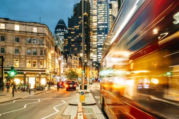 Motion-blurring red double-decker bus navigating busy London streets at dusk, with the iconic Gherkin skyscraper in the background
