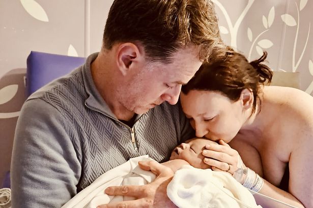 The image shows the parents holding their baby. Rhodri looks at the child solemnly while Emily kisses the baby's head. The baby is wrapped in a white towel