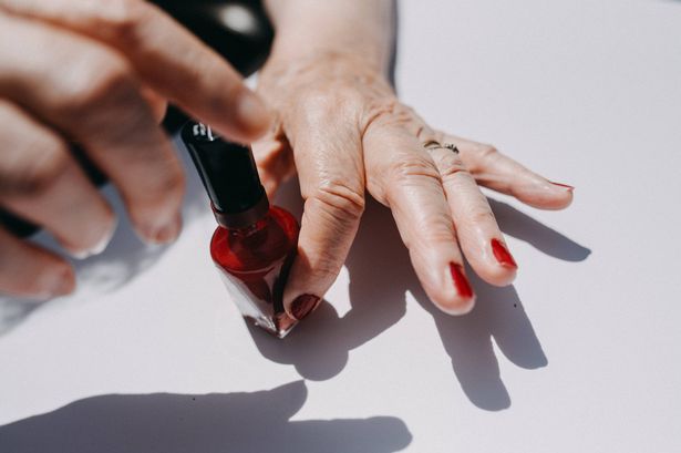A person is applying a bright red nail polish to their nails, with the bottle prominently displayed in the foreground. The well-lit scene emphasizes the contrast between the vibrant polish and the natural tones of the skin.