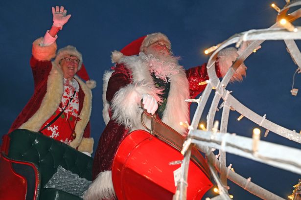 A person dressed as Santa Claus. He is dressed holding a metal rail, and woman in Christmas reds is standing behind him waving.