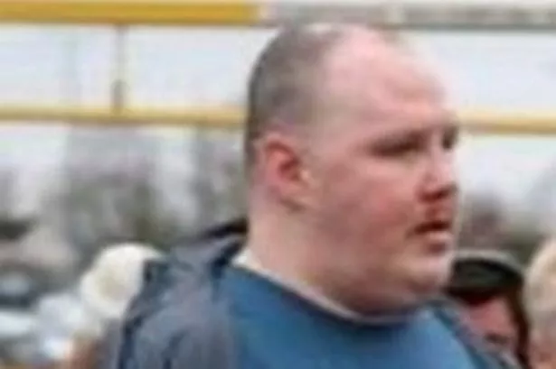 An image of a balding man in a crowd, wearing a jacket over a blue top with a logo.