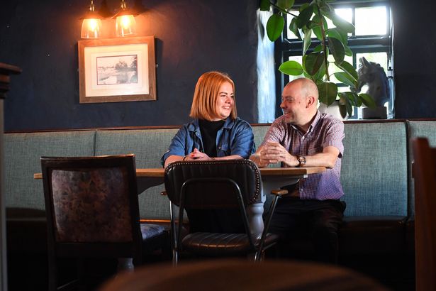 The image shows the two - Monika with red hair and wearing a denim jacket and Nathan with a bald head and wearing a smart collared shirt - chatting and laughing together 