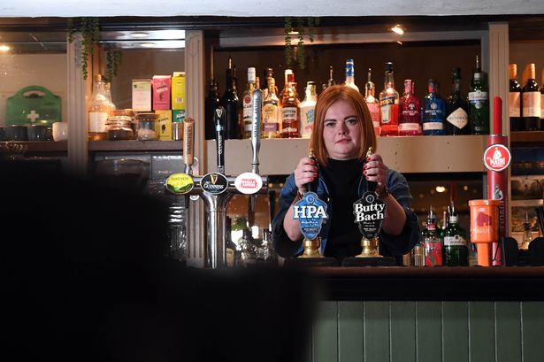 The image shows Monika, with red hair, fair skin and denim jacket, posing behind the bar with her hands on the kegs