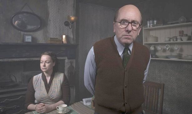 A man in glasses stands as a woman sits at a table